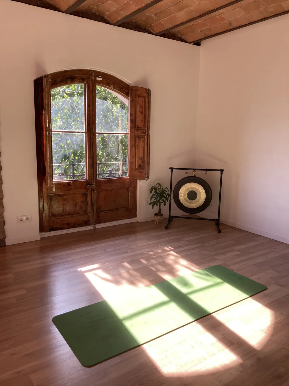 Sunlit corner of the Shala with an arched shuttered window, a gong and a mat on the wooden floor